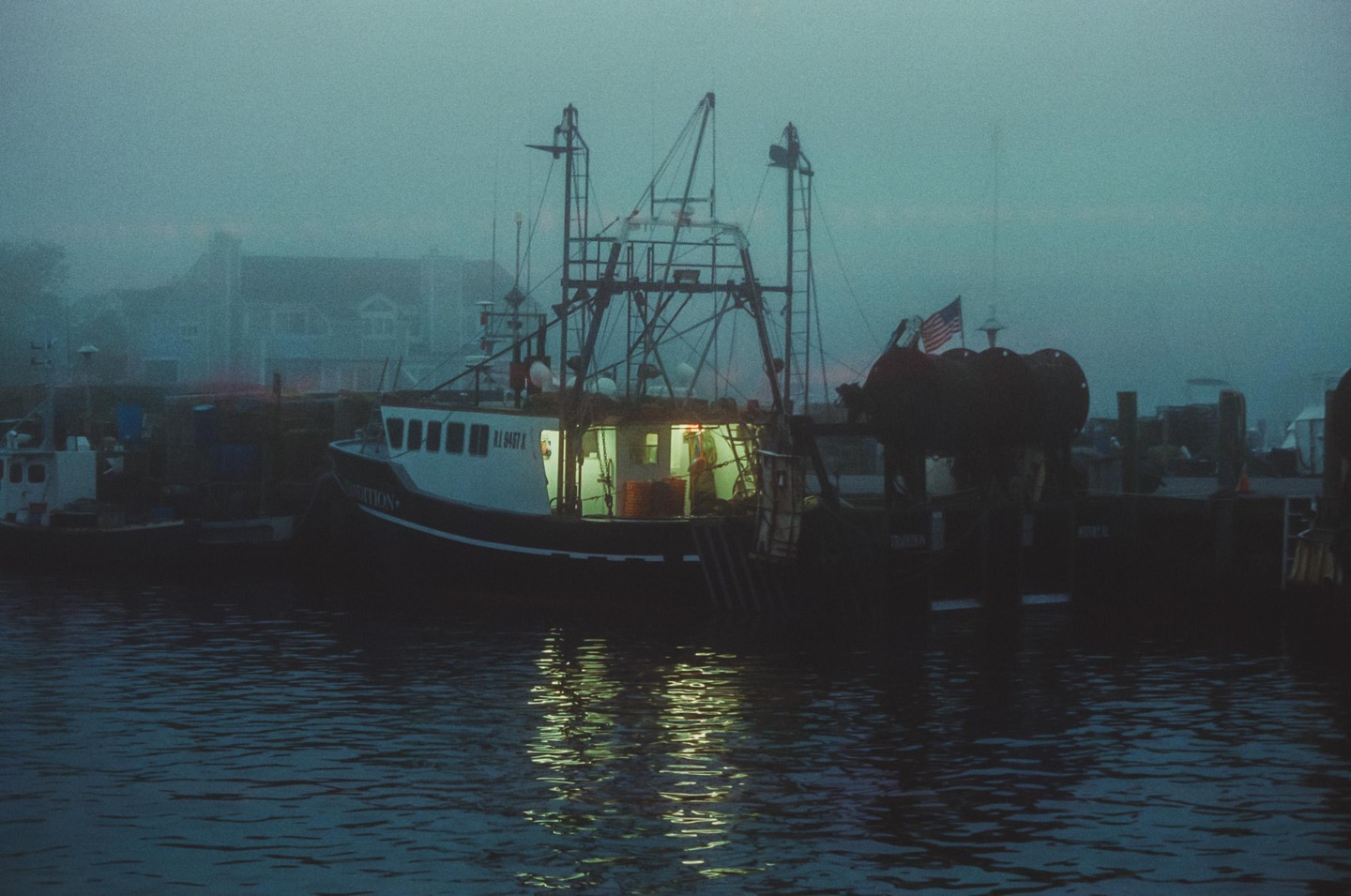 A fishing boat with illuminated windows is docked in a harbor on a foggy day.