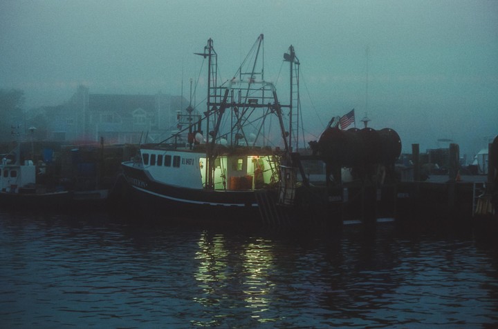 A fishing boat with illuminated windows is docked in a harbor on a foggy day.