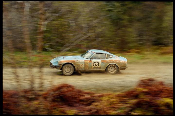 A light blue rally car with the number 53 on its side speeds along a dirt road, kicking up dust.