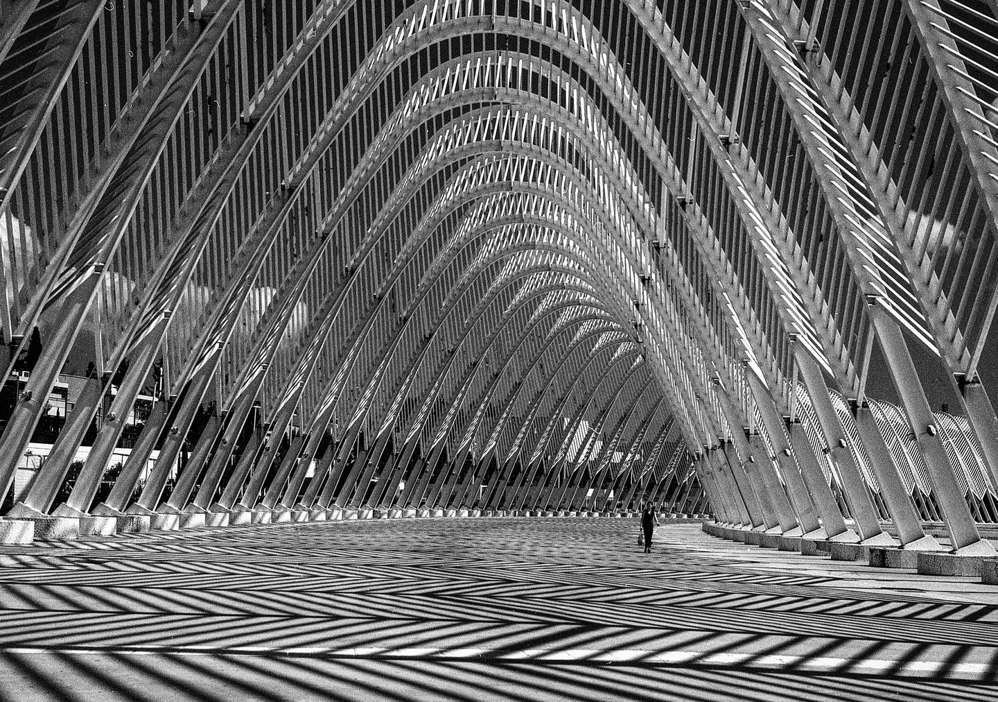 A lone person walks through a modern architectural structure with repeating arches and strong shadows on the ground.