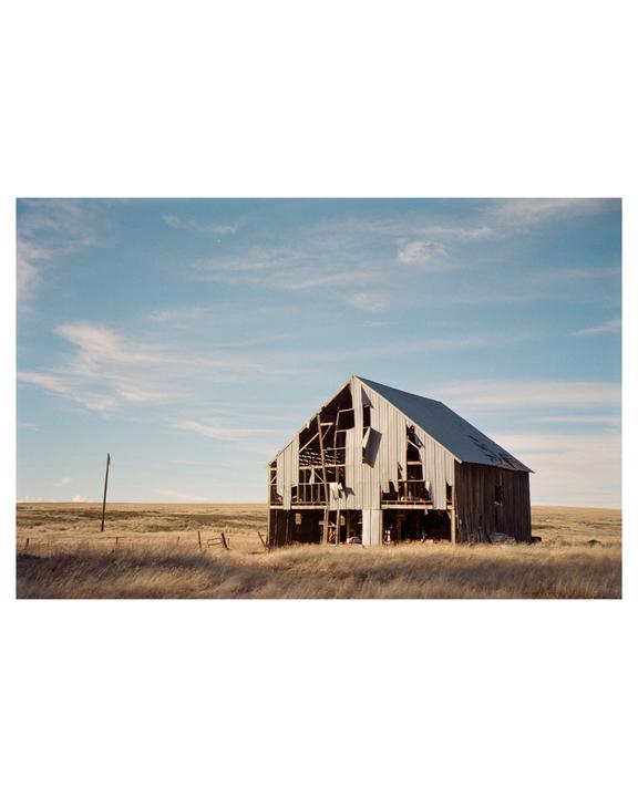 An abandoned barn with a partially collapsed roof stands in a field of dry grass under a blue sky.