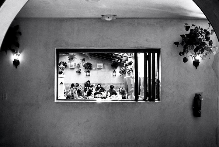A group of people are visible through a window, sitting at tables in a restaurant.