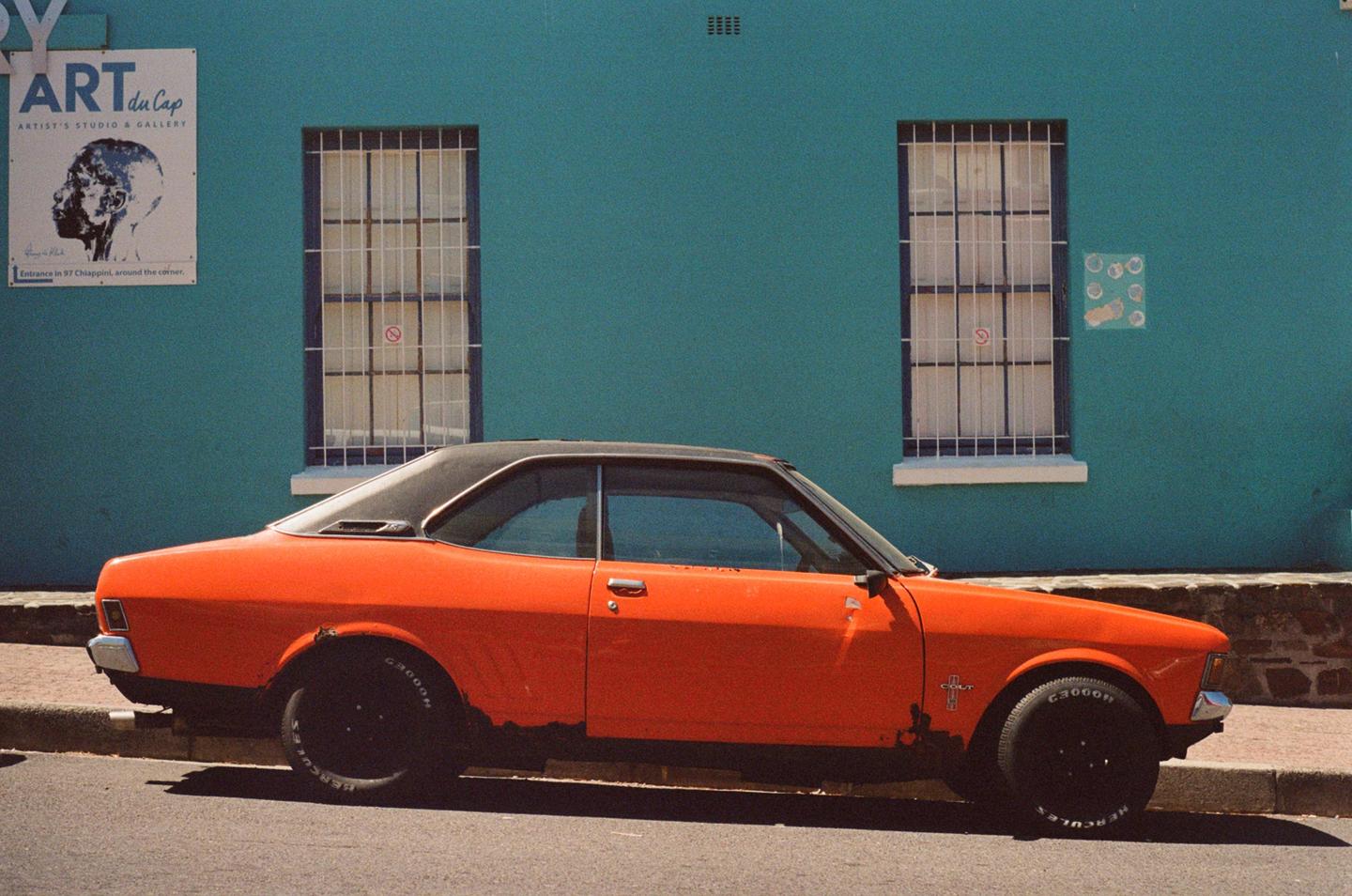 An orange car with a black roof is parked on a street next to a teal building with two windows.