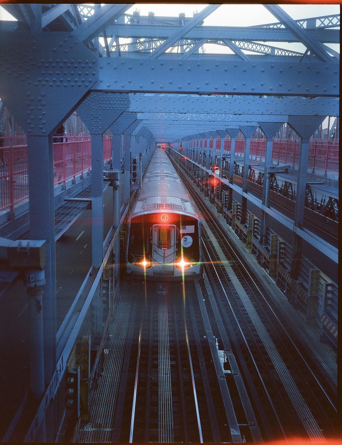 A subway train with illuminated headlights travels on tracks under a metal bridge structure.