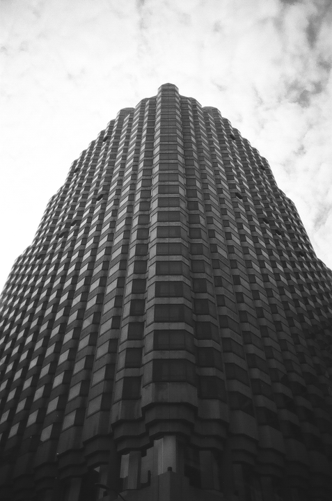 A tall building with a textured facade rises towards a cloudy sky.