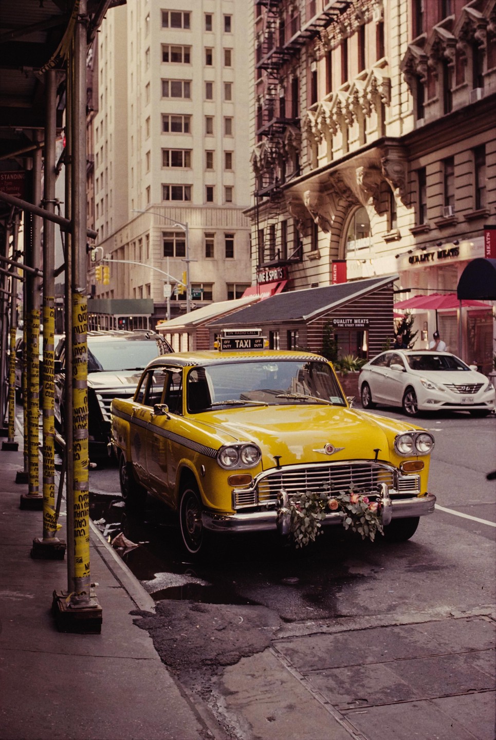 A yellow taxi cab with flowers on its bumper is parked on a city street next to scaffolding.