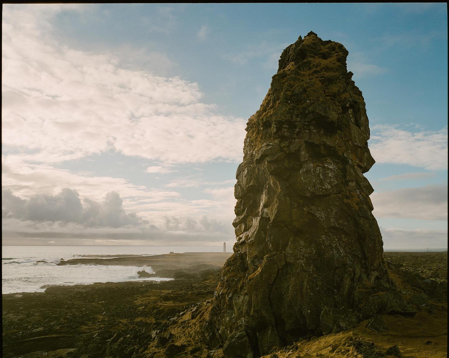 A large rock formation covered in moss stands prominently on a rocky coastline with a lighthouse in the distance under a cloudy sky.