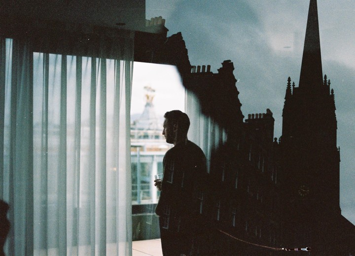 A man stands by a window with sheer curtains, looking out at a city view with a building reflection superimposed on the right.