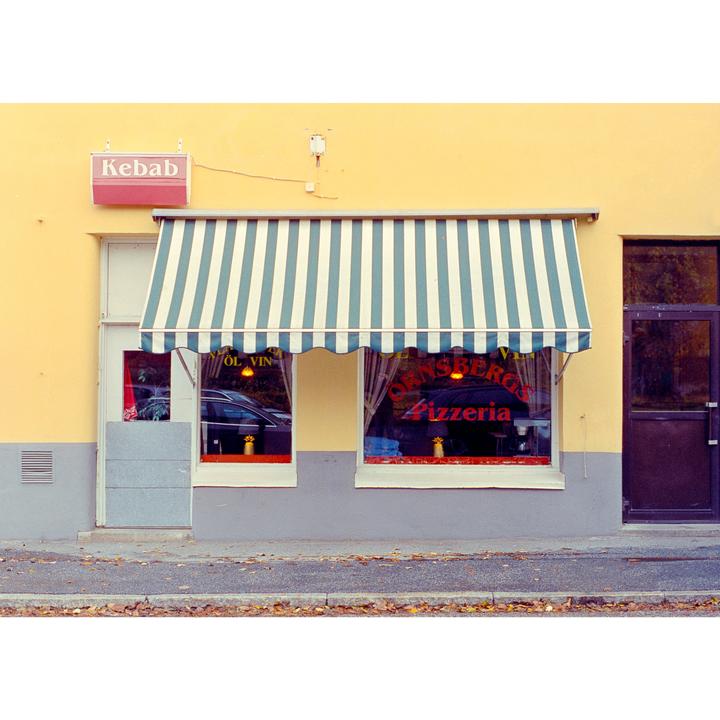 A yellow building with a striped awning over a pizzeria and a kebab sign is seen from across the street.