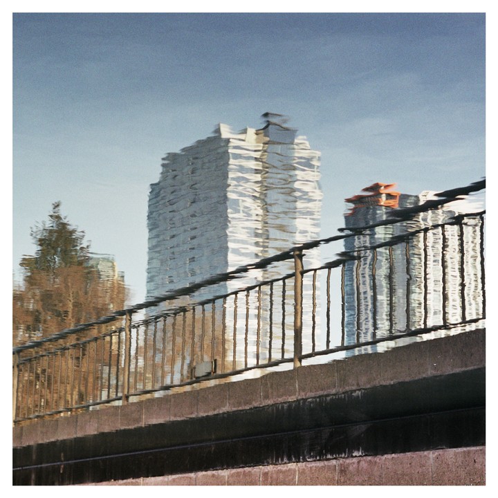 The reflection of two buildings and a tree are visible in rippling water behind a metal railing and concrete wall.