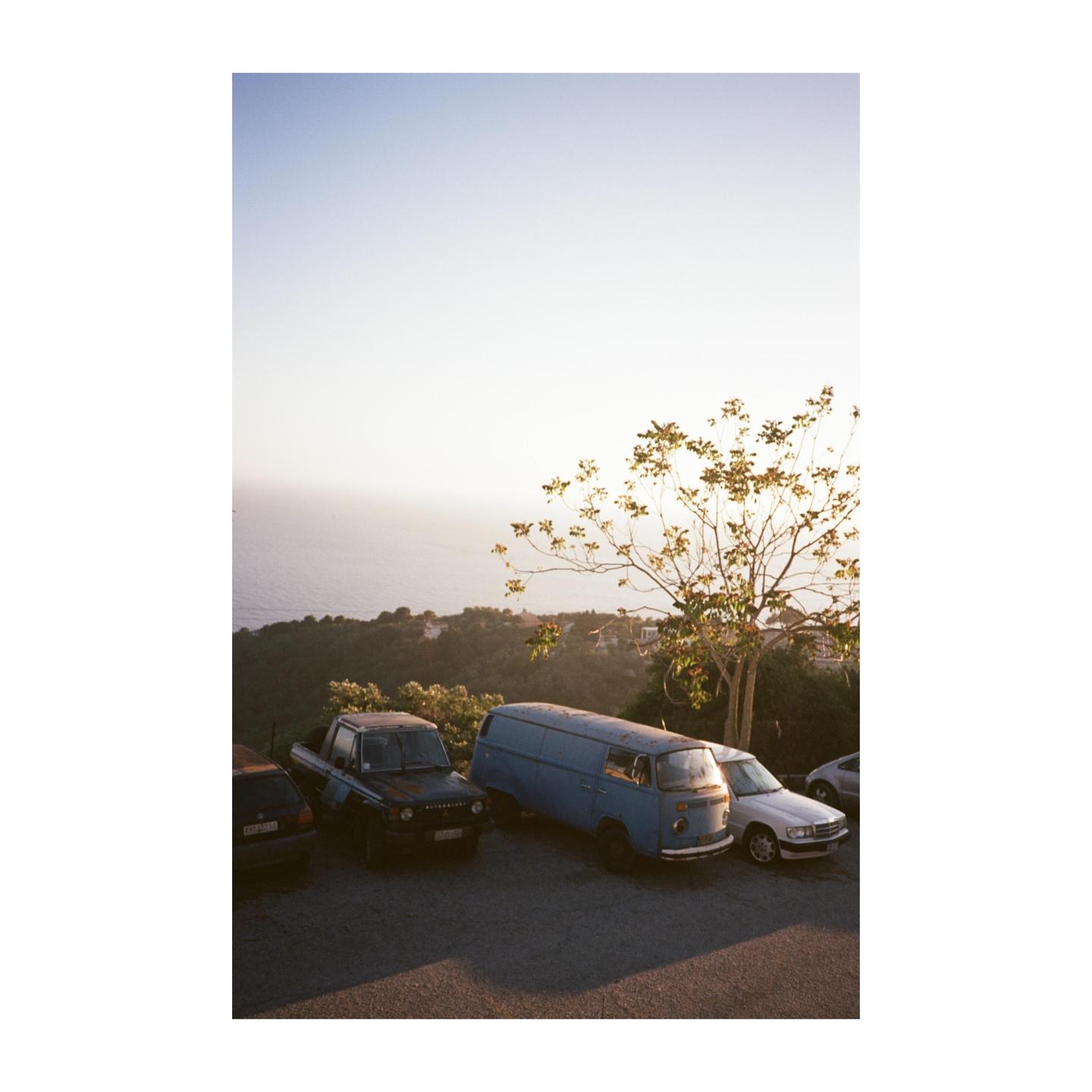 Several vehicles are parked on a paved area overlooking the sea and a tree with sparse foliage.