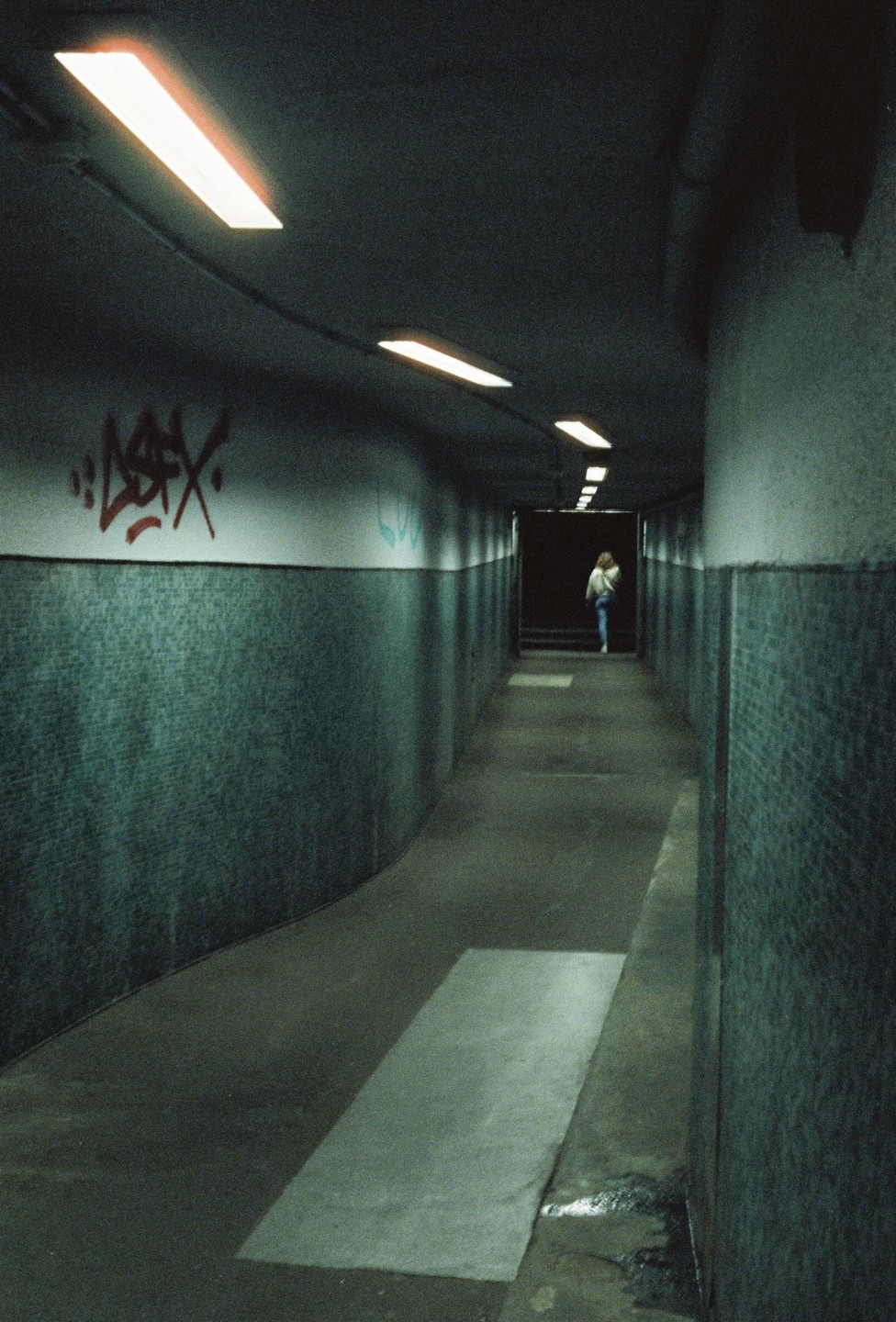 A person walks away down a long, dark, curving tunnel with fluorescent lights on the ceiling and graffiti on the wall.