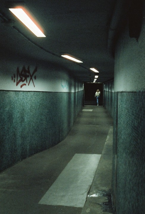 A person walks away down a long, dark, curving tunnel with fluorescent lights on the ceiling and graffiti on the wall.