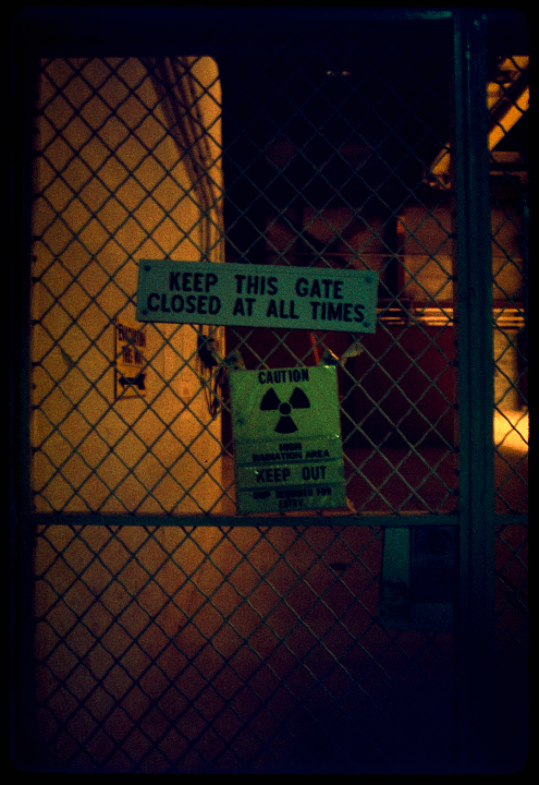 A chain link fence with two signs, one reading "KEEP THIS GATE CLOSED AT ALL TIMES" and another "CAUTION HIGH RADIATION AREA KEEP OUT", is visible at night.