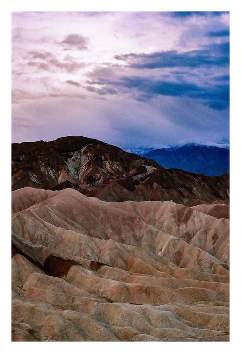 A dramatic sky with purple and blue clouds hangs over a desert landscape with rolling hills and mountains.