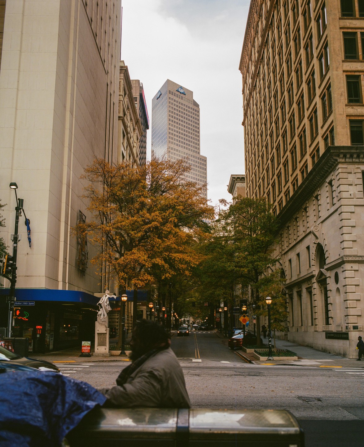 A man sits in the foreground of a street scene with tall buildings and autumn trees.