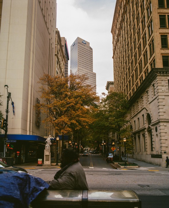 A man sits in the foreground of a street scene with tall buildings and autumn trees.