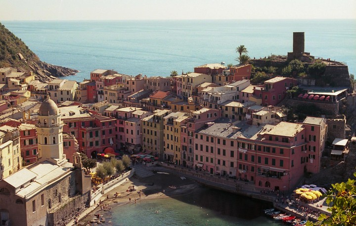 A colorful village with a church and a castle tower sits on a hillside overlooking the sea.