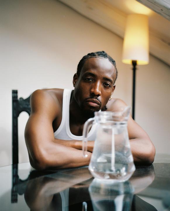 A man with braided hair sits at a table with a pitcher of water in front of him.