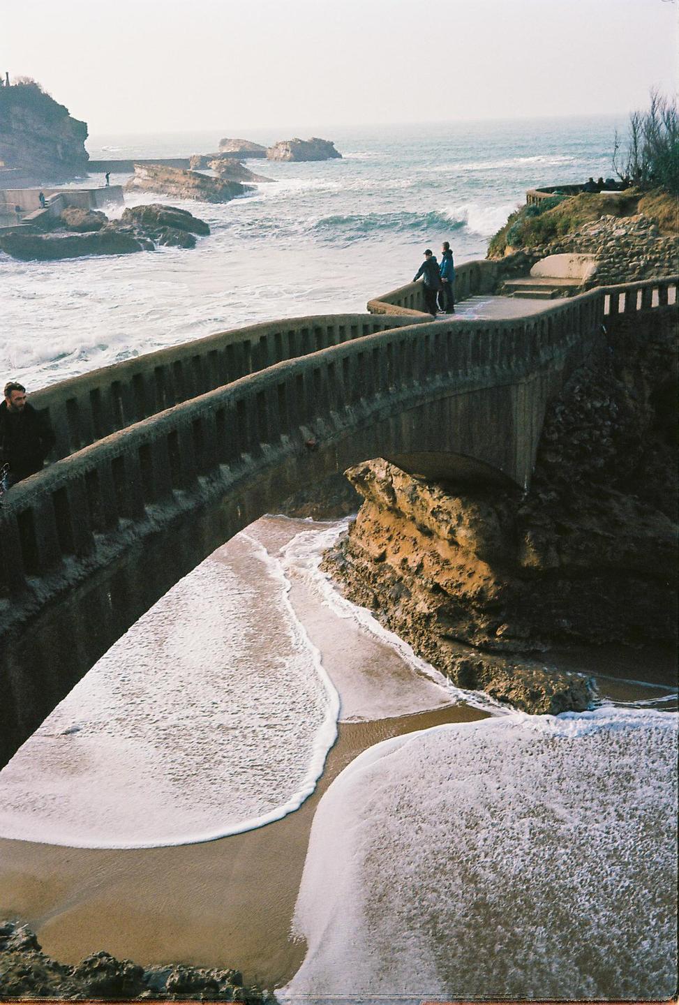 A concrete bridge curves along a rocky coastline with waves crashing on the shore and a sandy beach below.