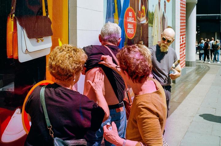 Two women apply cream to a man's back on a sunny street.