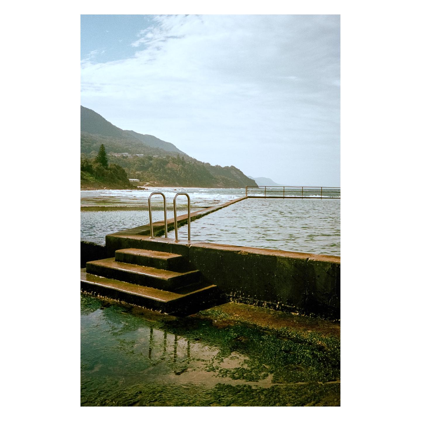 A concrete rock pool with steps and handrails sits beside a beach with waves, with mountains in the background under a cloudy sky.