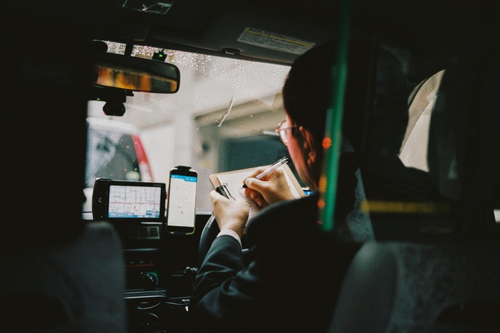 A man in a suit writes on a clipboard while sitting in the driver's seat of a taxi.