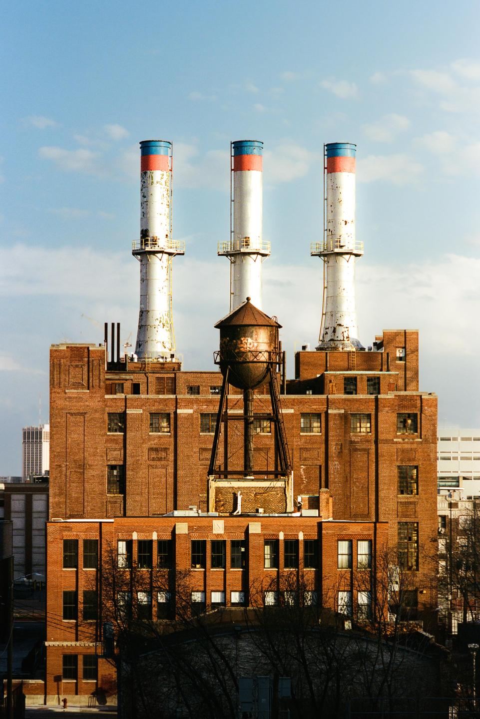 Three tall industrial smokestacks with red and blue tops rise above a brick building with a water tower on its roof.