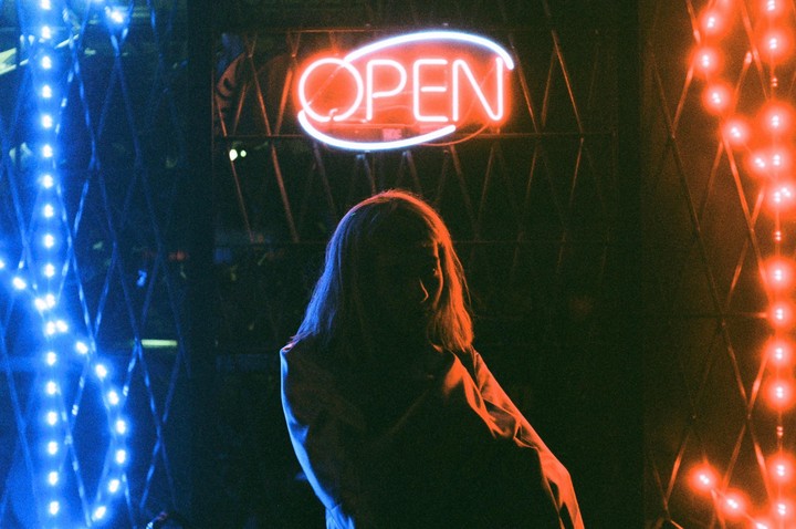 A woman stands in front of an "OPEN" neon sign, illuminated by blue and red lights.