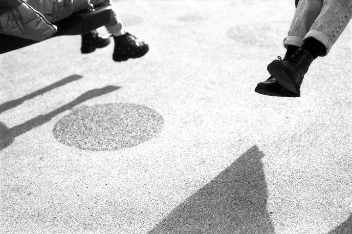 Two people's legs and feet are visible above a textured ground with shadows.