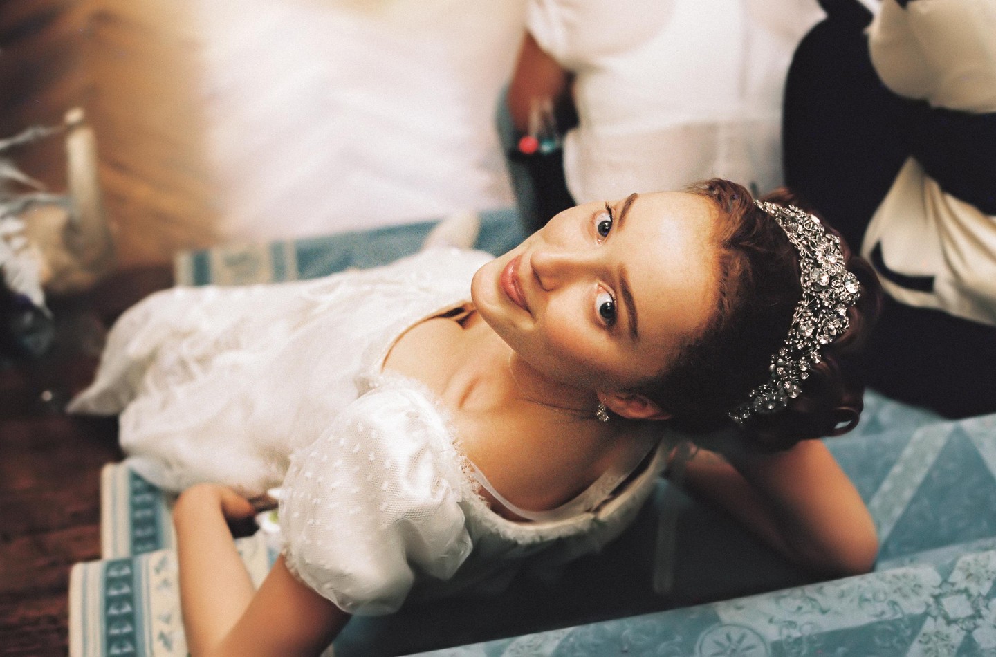 A woman in a white dress and a jeweled headband is looking up at the camera while lying on a patterned blue surface.