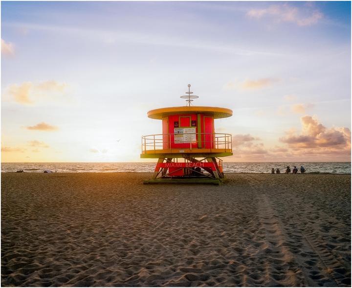 A colorful lifeguard stand stands on a sandy beach with the ocean and a sunset in the background.