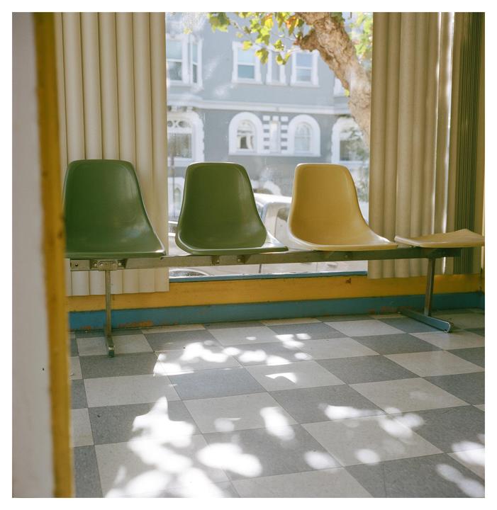 Three empty chairs are lined up in a waiting room with a checkered floor and a window looking out onto a building and tree.