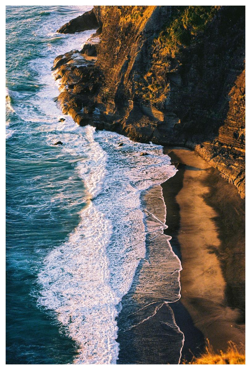 Waves crash against a rocky cliff and sandy beach during golden hour.