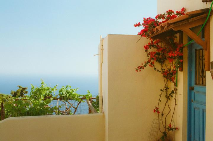 Red bougainvillea flowers grow over a blue door and a tiled awning, with the sea visible in the distance.