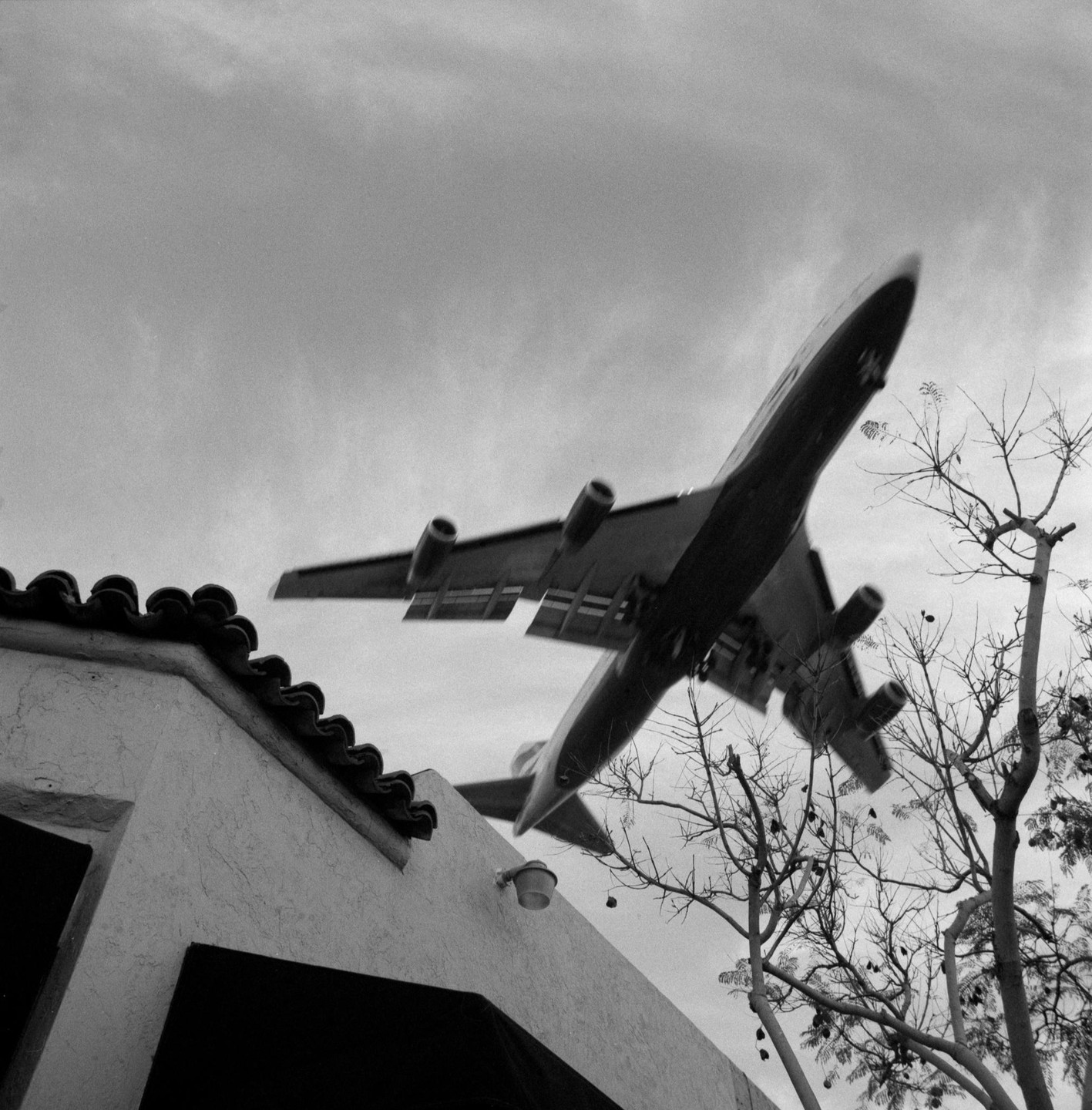 A large airplane flies low over a building with a tiled roof and bare trees.