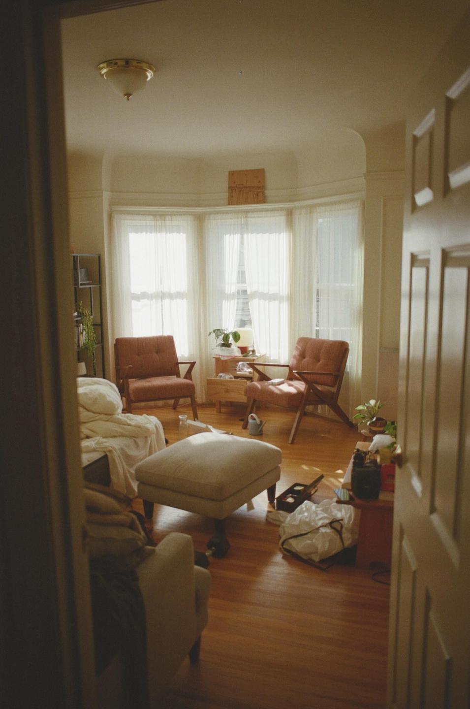 Sunlight streams through sheer curtains into a living room with two armchairs, an ottoman, and a bed.