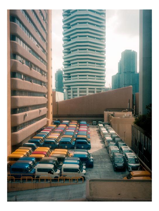 A parking lot full of colorful kei cars is nestled between tall buildings.