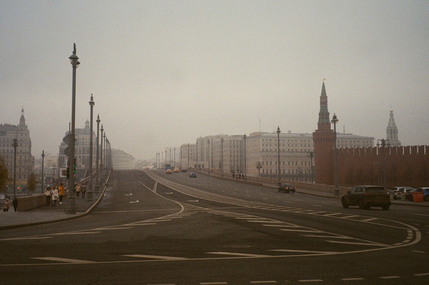 A wide street with cars and pedestrians is visible under a hazy sky, with historic buildings and a red brick wall lining the road.