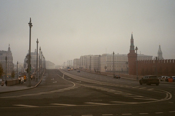 A wide street with cars and pedestrians is visible under a hazy sky, with historic buildings and a red brick wall lining the road.