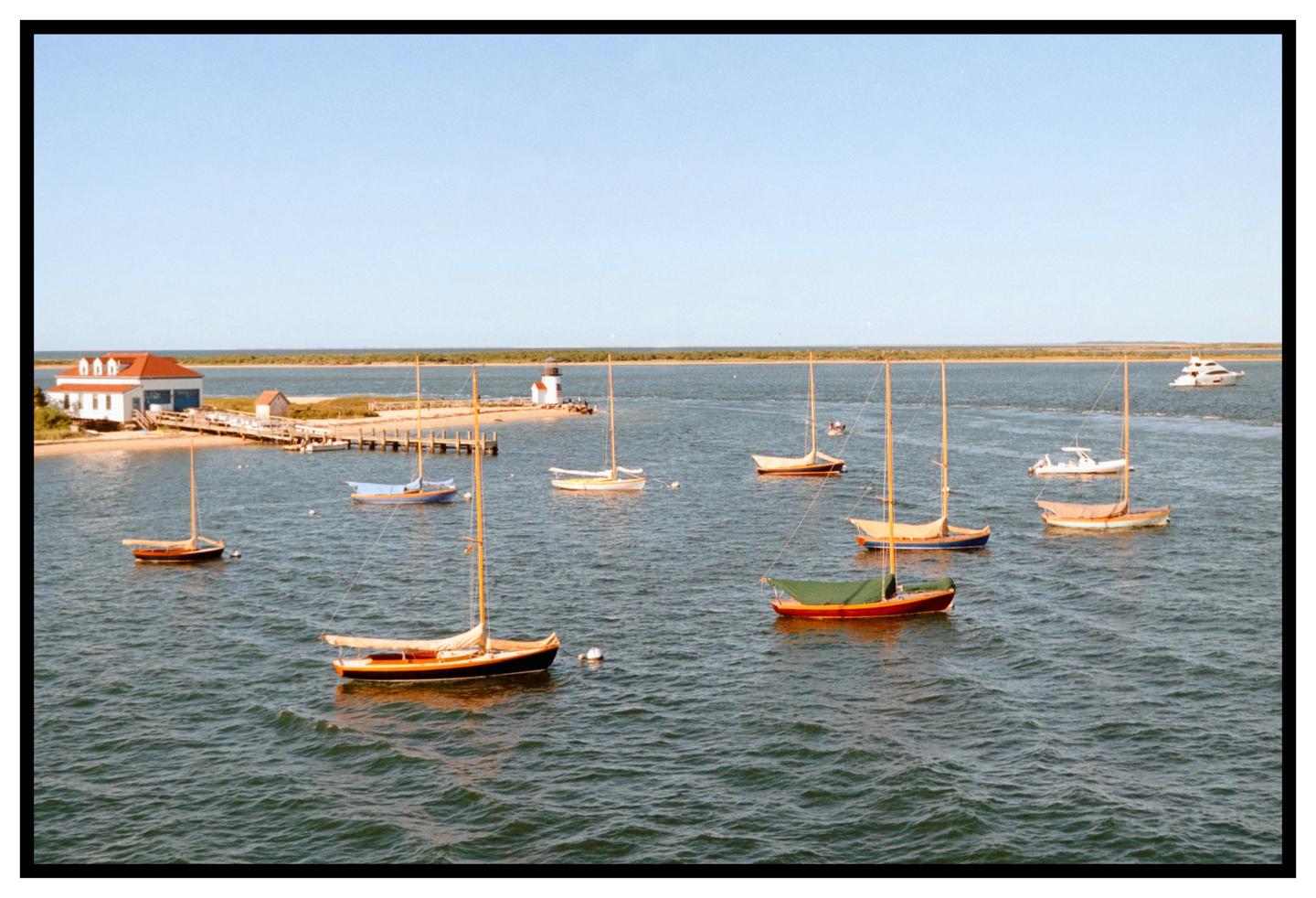 Numerous sailboats are moored in a calm body of water with a small building and lighthouse visible on the distant shore.