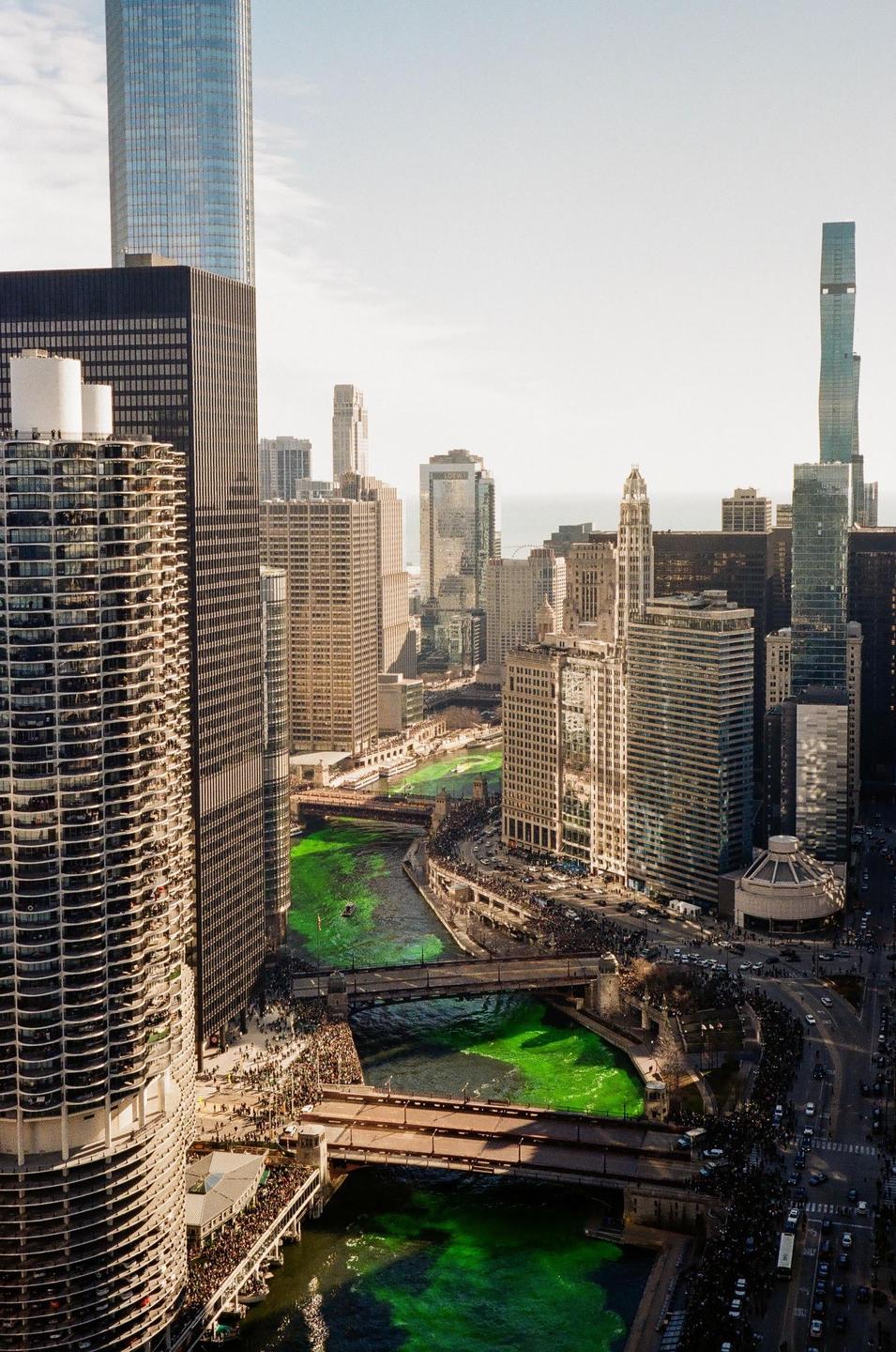 An aerial view shows a city with a river dyed green, surrounded by tall buildings and crowds of people.