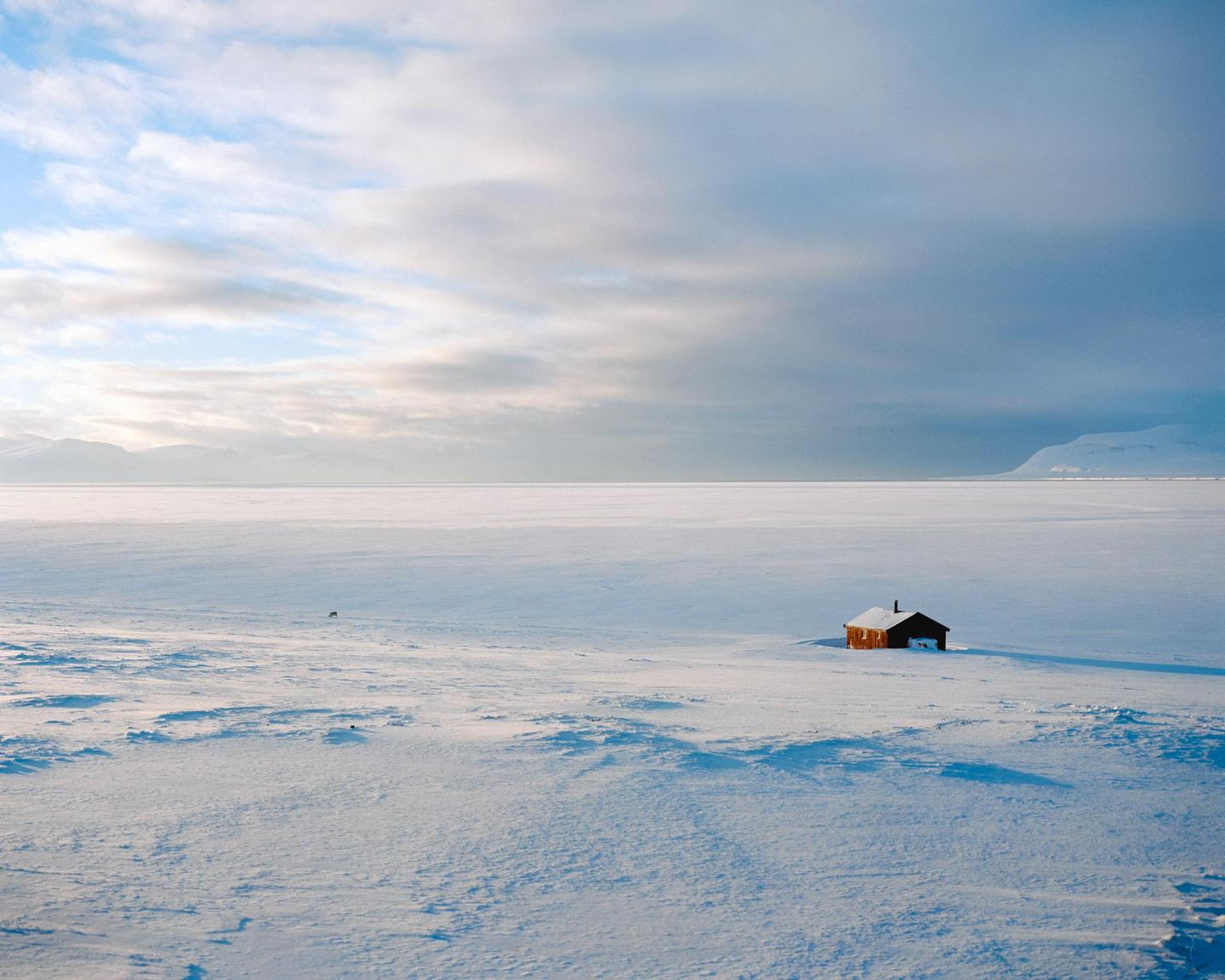 A small, dark cabin sits alone in a vast, snow-covered landscape under a cloudy sky.