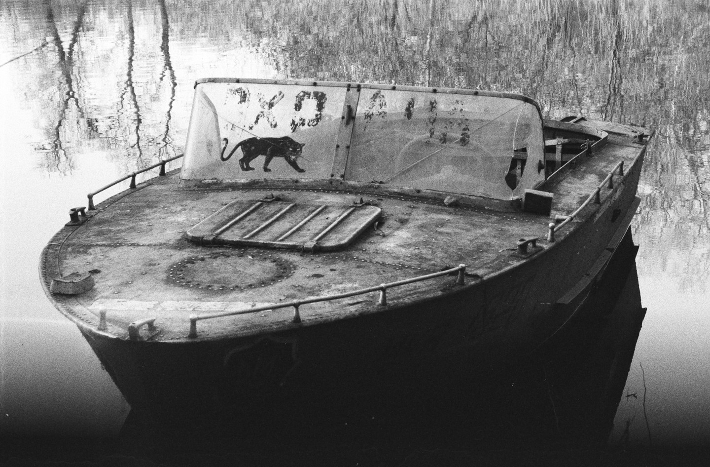 An abandoned boat with a panther decal on its windshield floats on water that reflects trees.