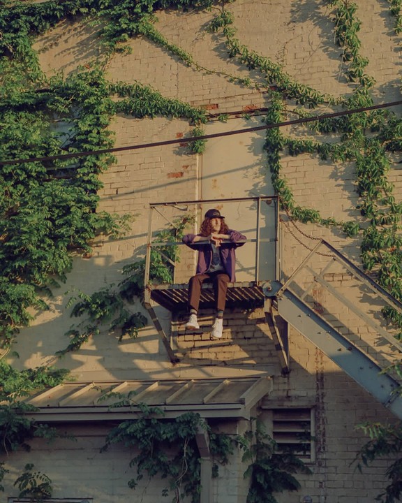 A person with long hair and a cap sits on a fire escape platform against a brick wall covered in ivy.