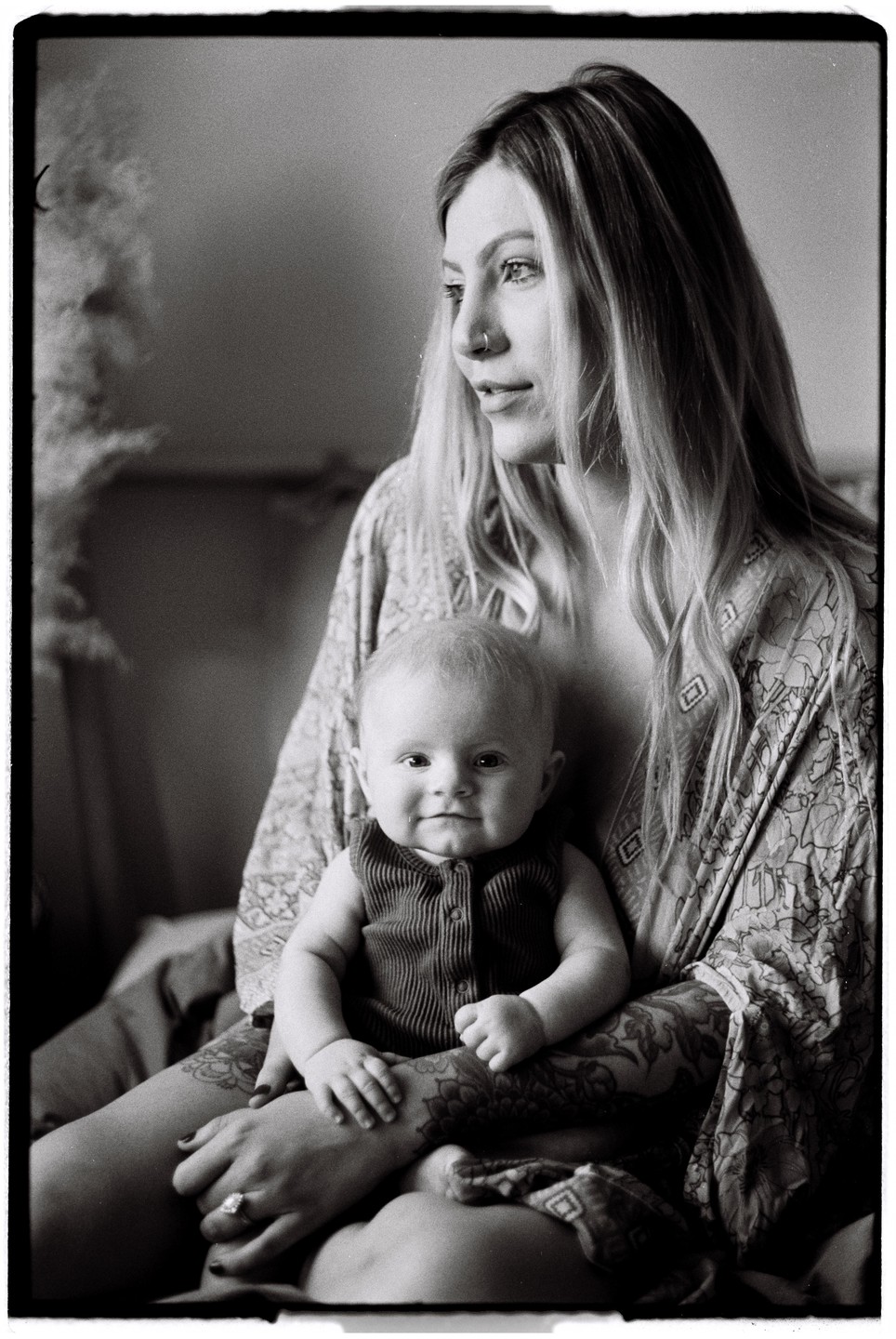 A woman with long hair and a baby on her lap are sitting in window light.