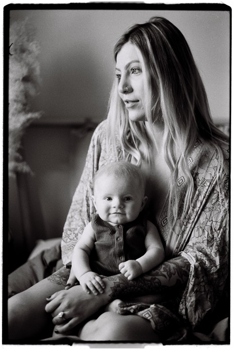 A woman with long hair and a baby on her lap are sitting in window light.
