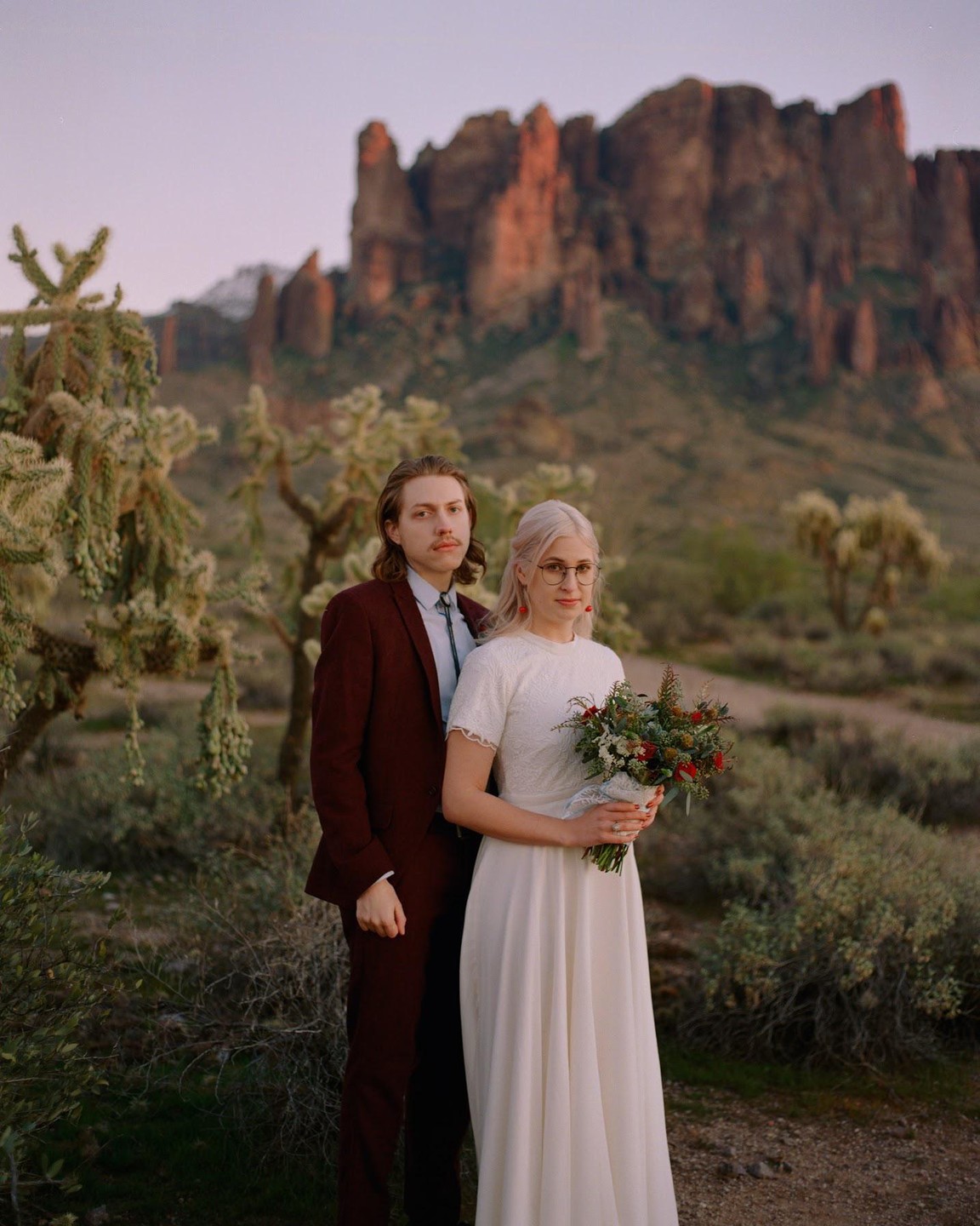 A couple stands in a desert landscape with mountains in the background.