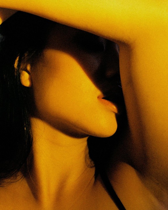 A woman's face is partially obscured by shadow, with her lips slightly parted and her tongue visible.