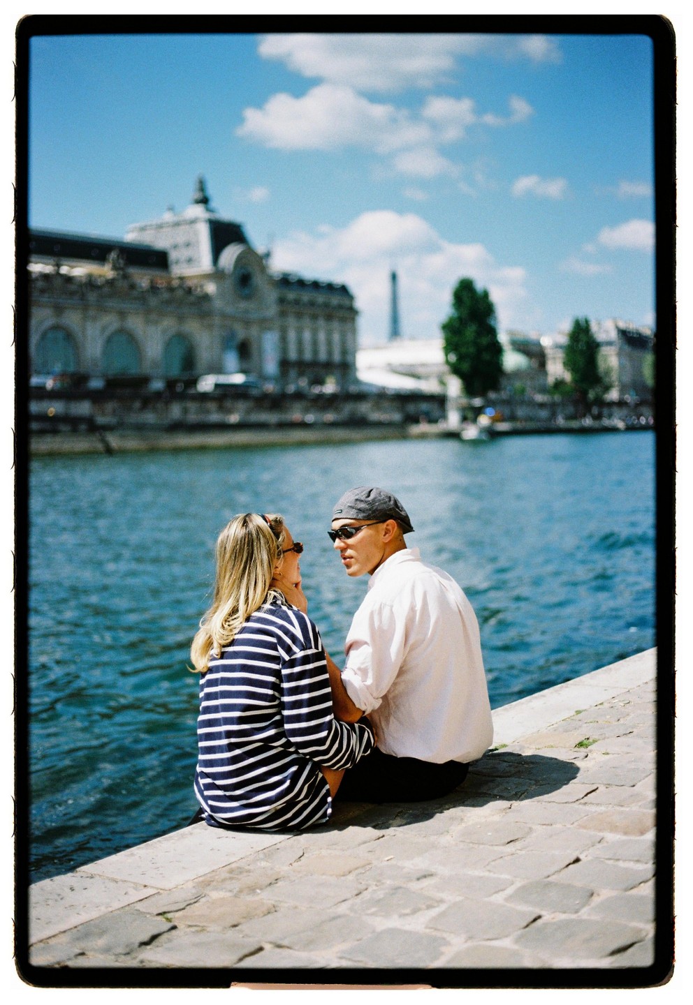 A couple sits by the water with a large building and the Eiffel Tower in the background.